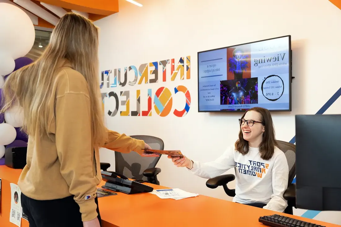 Photo of Gilmore sitting behind a desk at the Intercultural Collective, smiling and handing a student.